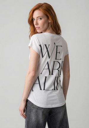 Rothaarige Frau trägt ein weißes T-Shirt mit großem schwarzem Schriftzug "WE ARE ALIVE" auf der Rückseite, kombiniert mit grau verfärbten Jeans.