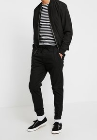 Czarna kurtka bomberka nałożona na koszulkę w paski, w zestawieniu z czarnymi zwężającymi się spodniami i czarnymi trampkami. Stylizacja charakteryzuje się luźnym krojem i elastycznymi detalami.