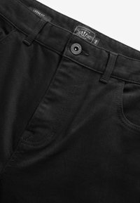 Sorte løstsiddende bukser med synlige knapper, bæltestropper, forlomme og et mærke med størrelse 34R og mærkeoplysninger.