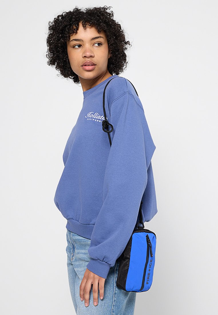 Blauer Pullover mit gerippten Bündchen, getragen mit hoch taillierten Jeans. Eine kleine schwarz-blaue Umhängetasche hängt von der Schulter und hat einen Reißverschluss.
