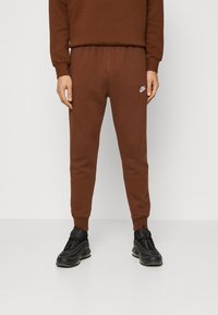 Muž oblečený v hnědých teplákových kalhotách Nike s logem na stehně, černých teniskách, stojící proti jednobarevnému bílému pozadí.