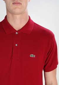 Polo rouge en tissu texturé, avec un col, deux boutons et un petit logo crocodile vert brodé sur le côté gauche de la poitrine.