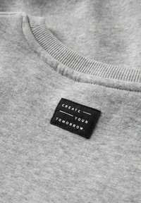 Sudadera gris con cuello redondo acanalado y etiqueta rectangular negra cosida que dice "CREATE YOUR TOMORROW." en texto blanco.
