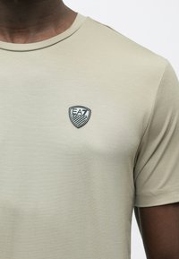 Bézs rövid ujjú póló puha, sima anyagból. Bal oldalon a mellrészen fekete EA7 logófolt, és kerek nyakkivágás jellemzi.