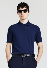 Polo shirt blu navy con tessuto testurizzato, colletto a tre bottoni e maniche corte. Indossato con pantaloni scuri e una cintura nera.