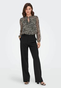 Blouse en tulle imprimé léopard avec un col froncé, associée à un pantalon noir taille haute. Le mannequin arbore des vagues naturelles et des accessoires minimalistes.