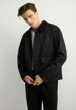 Młody mężczyzna w czarnej zapinanej na guziki kurtce na białej koszuli, poprawiający suwak kurtki, stojący na jednolitym jasnym tle.