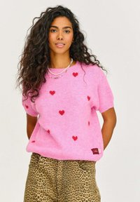 Ružový sveter s krátkymi rukávmi vyrobený z mäkkého materiálu, s rozptýlenými červenými srdiečkami a malou brandovou etiketou na leme. V kombinácii s nohavicami s leopardím vzorom.