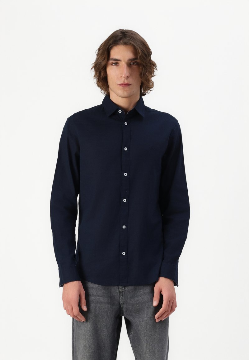Camisa de botonadura en azul marino con cuello, mangas largas y botones blancos, combinada con unos jeans de mezclilla gris.