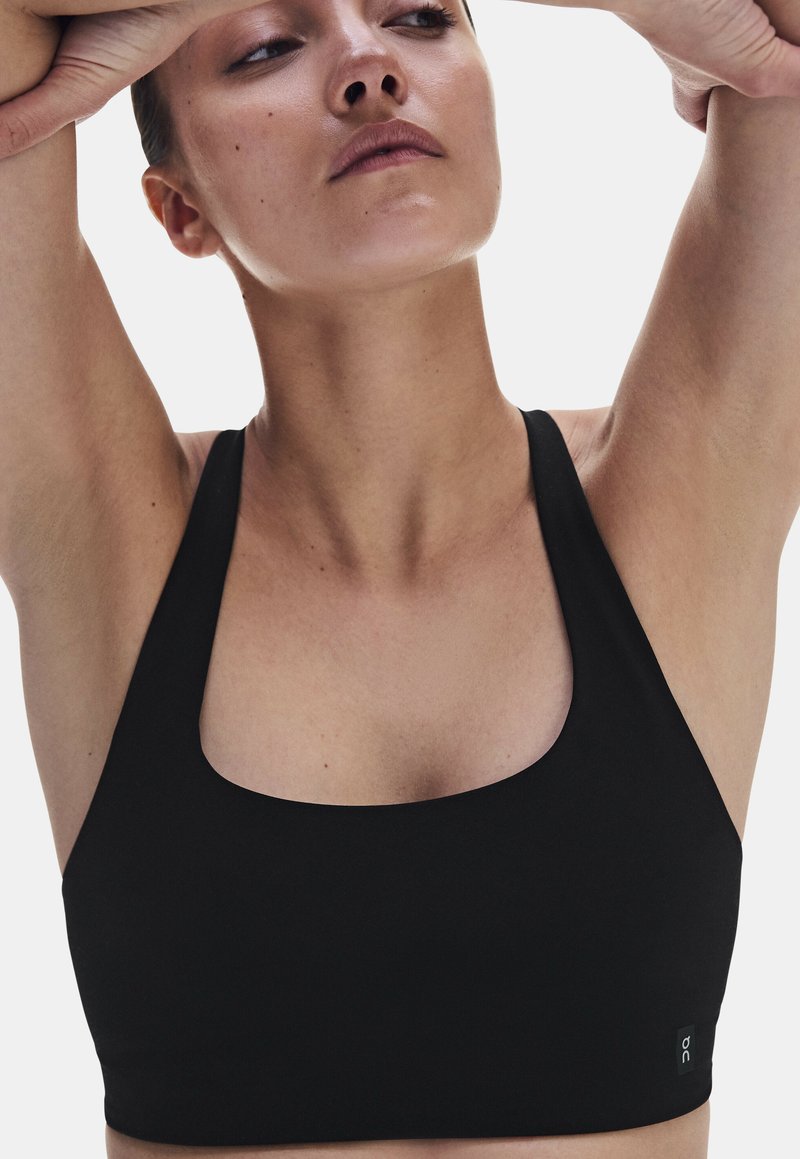 Femme portant une brassière de sport noire, les bras levés au-dessus de la tête, regardant légèrement vers le haut sur un fond blanc uni.