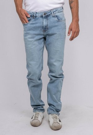 Homme portant un jean bleu clair, des baskets blanches avec une étoile noire, et un t-shirt blanc, une main dans la poche, sur un fond uni.