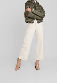 Olivgrüner gesteppter Puffer-Jacke mit Kapuze, kombiniert mit cremefarbenen weiten Hosen und beigen, glatten, hochhackigen Stiefeletten.