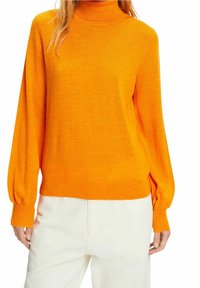 Pull orange à col montant, manches longues et coupe décontractée. Texture tricotée ; poignets et ourlet côtelés. Associé à un pantalon blanc.