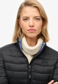 Czarna kurtka puchowa o pikowanej fakturze, z wysokim kołnierzem i zapięciem na zamek, noszona na kremowym swetrze z golfem o miękkiej, puszystej teksturze.