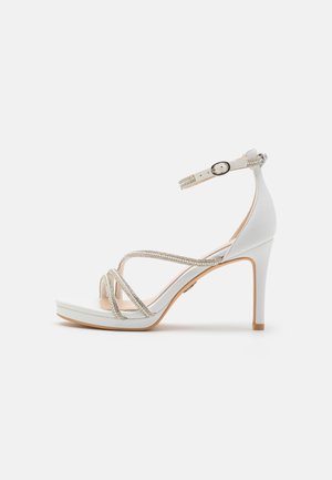 Λευκά πέδιλα με ψηλό τακούνι και πλατφόρμα, με σταυρωτά λουριά διακοσμημένα με ασημί ριχνοστάτες και ρυθμιζόμενο λουρί στον αστράγαλο.
