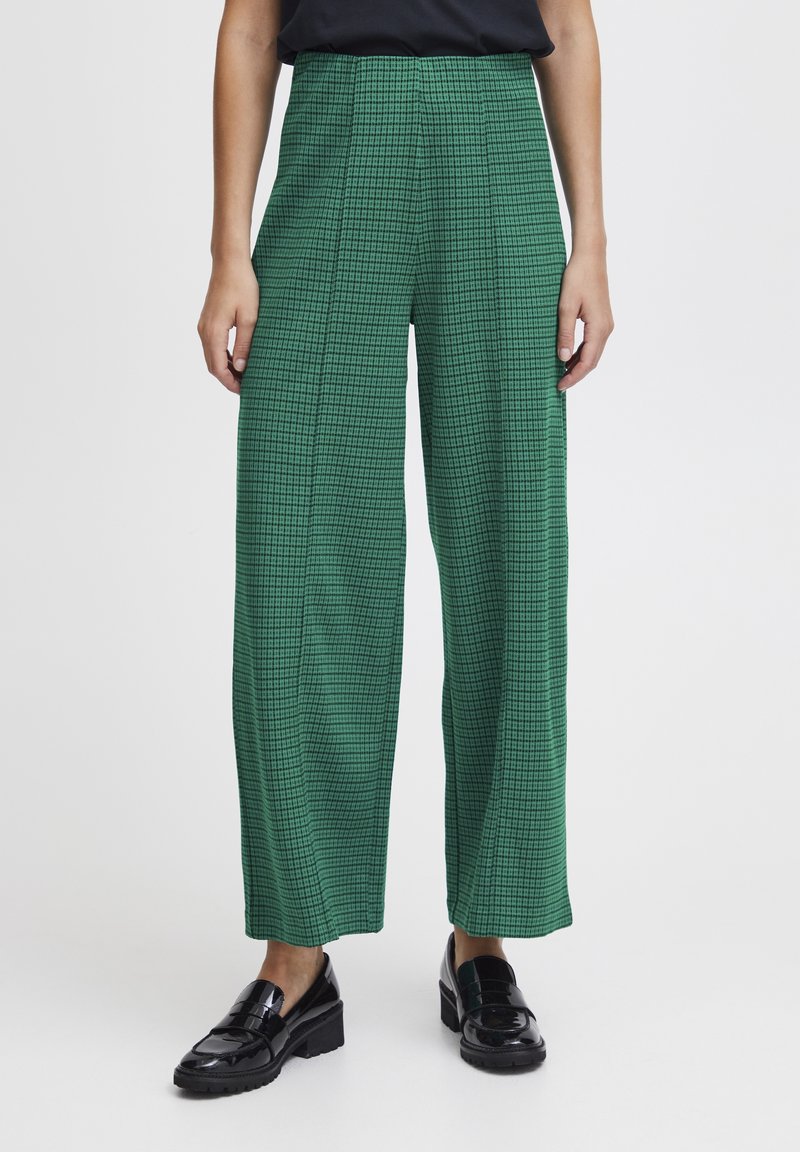 Femme portant un pantalon large vert à carreaux taille haute et des mocassins en cuir noirs, debout sur un fond uni.
