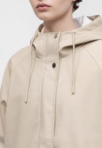 Gros plan d'une personne portant une veste à capuche beige avec cordons et boutons-pression métalliques sur la fermeture avant.