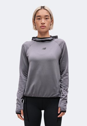 Jeune personne aux cheveux blonds courts portant un sweat à capuche gris à manches longues New Balance et des leggings noirs sur un fond uni.