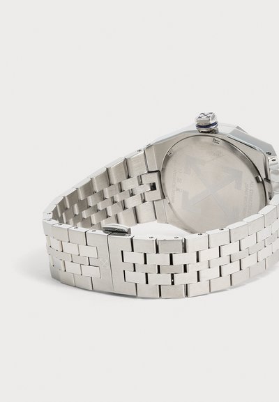 Montre-bracelet en acier inoxydable avec bracelet à maillons, montrant une boucle fermée et un fond gravé avec logo et détails de résistance à l'eau.