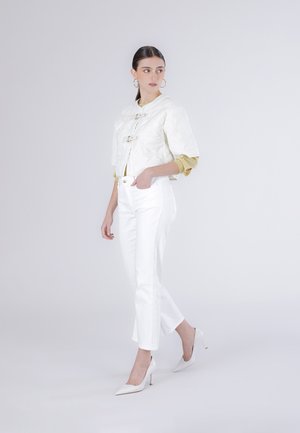 Kvinde stående iført hvid quiltet kort jakke, gul top, hvide højtaljede bukser og hvide højhælede sko på en ensfarvet baggrund.