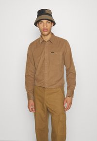 Ruda korduroy mygtukinė marškiniai su krūtine kišene, derinami su rudais cargo džinsais. Dviejų tonų juodas ir rudas kibirinis skrybėlė užbaigia derinį.