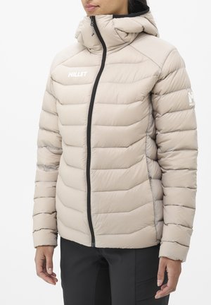 Femme portant une veste matelassée beige avec capuche, fermeture éclair noire, et logos "Millet" sur la poitrine et la manche.
