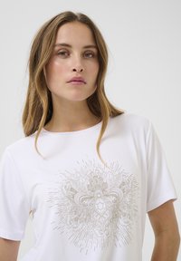 T-shirt blanc à manches courtes avec un design graphique doré détaillé au centre, présentant des motifs complexes et des lignes rayonnantes.