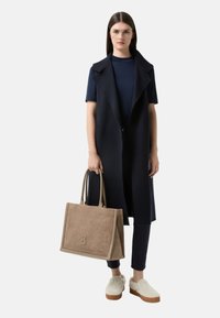 Námořnický dlouhý vest over tmavým topem, v kombinaci s černými kalhotami. Drží béžovou tašku s embosovaným logem a má na sobě béžové slip-on boty.