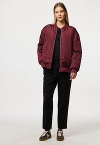 Burgundy bomber jacket with a smooth finish, ribbed cuffs, and a front zip, paired with black trousers and patterned sneakers.