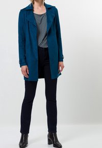 Türkisfarbener Wildleder-Trenchcoat mit breiten Revers, kombiniert mit einem gemusterten Oberteil, schwarzen Skinny-Jeans und Ankle Boots. Glatte Textur, mittellanges Design.