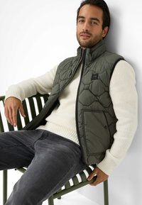 Gepatchte grüne Weste mit hohem Kragen über einem klobigen weißen Pullover, kombiniert mit dunkelgrauen Jeans und sitzt auf einem grünen Stuhl.