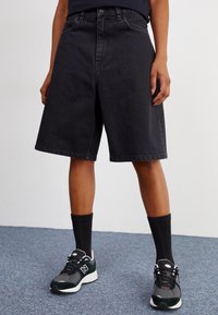 Svarte denimshorts med bred passform, med høy midje og frontlommer. Kombinert med svarte ribbestrikkede sokker og grå joggesko.