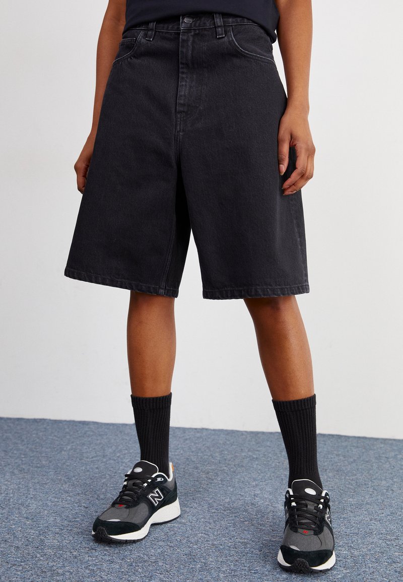 Svarte denimshorts med bred passform, med høy midje og frontlommer. Kombinert med svarte ribbestrikkede sokker og grå joggesko.