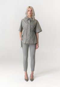 Lichtgrijs, kort mouwen overhemd met kraag en een taillekoord, gecombineerd met nauwsluitende grijze broek en hakken, tegen een effen achtergrond.