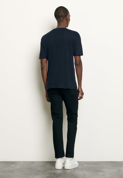 Tricou cu mânecă scurtă de culoare bleumarin, cu guler rotund, purtat împreună cu pantaloni negri și adidași albi. Țesătura pare ușoară și texturată.