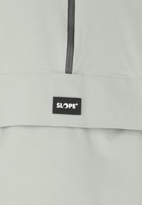 Lysegrå jakke med halvlynlås, prydet med et sort SLOPE® logo-patch på brystet og en glat, mat overflade.