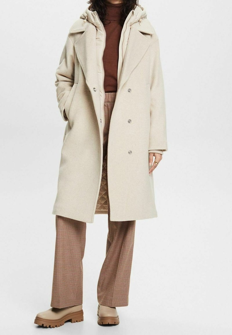 Abrigo de mezcla de lana color beige con una capa interior acolchada, grandes solapas y cierre de doble botonadura. Combinado con pantalones de cuadros marrones y botas beige.