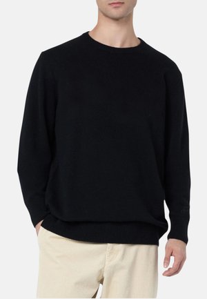 Uomo con maglione nero semplice con scollo tondo e pantaloni beige chiari, mani nelle tasche, su sfondo semplice.