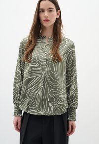 Blusa verde con un motivo astratto bianco, maniche lunghe con polsini arricciati e patta con bottoni, realizzata in tessuto liscio.