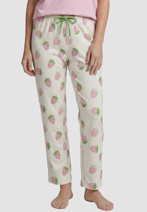 Vrouw draagt witte pyjamabroek met roze aardbeienprint en een groene trekkoord, gecombineerd met een lichtroze top, blootsvoets staand.