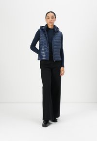Navy blue puffer vest, quilted design, zip-up front, attached hood, worn over a navy long-sleeve top, paired with black wide-leg pants and shoes.