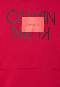 Sudadera roja con un destacado logo negro que dice "CALVIN KLEIN". La tela tiene una textura suave y hay una pequeña etiqueta.