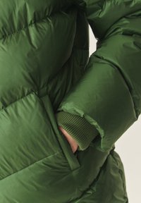 Giacca a vento verde con design trapuntato, polsini elasticizzati e tasca laterale. Il materiale ha una texture liscia e lucida.