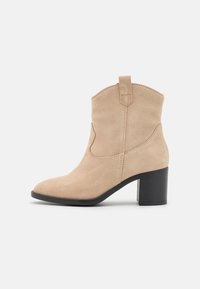 Krémové semišové kotníkové boty s špičatou špičkou a černým hromadným podpatkem. Disponují bočními švy a poutkem na holeně.