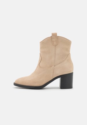 Beige suède enkellaars met een spitse neus en zwarte gestapelde hak. Voorzien van zijnaadaccenten en een lussen aan de schacht.