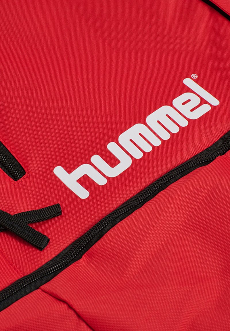 Mochila roja con un tejido duradero, que presenta un logotipo blanco prominente y acentos de cremallera negra. Superficie texturizada con costuras visibles.