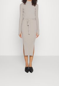 Robe en maille beige côtelée à manches longues avec un col montant et une taille ceinturée, présentant des fentes latérales et une longueur mi-mollet. Chaussures noires à enfiler visibles.