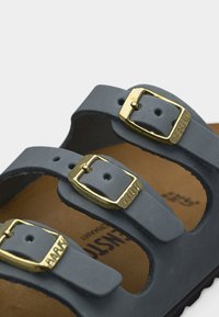 Μπλε λουριά σανδαλιών με τρεις ρυθμιζόμενες αγκράφες σε χρυσό τόνο πάνω σε ταμπά πέλμα και μαύρη σόλα, με μερική επωνυμία στο εσωτερικό.