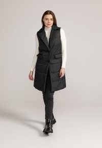 Zwarte gequiltte puffer vest met een sjaalkraag, knoopsluiting aan de voorkant en twee zijzakken, over een witte geribbelde trui en zwarte jeans.