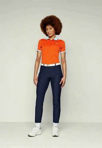 Γυναίκα με αφρο μαλλί, φορώντας κολάν πολο με κοντό μανίκι σε πορτοκαλί χρώμα, ναυτικές παντελόνες, άσπρη ζώνη, άσπρα παπούτσια, που στέκεται μπροστά σε απλό φόντο.